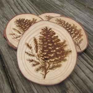 May include: Set of four wooden coasters with a natural wood grain. Each coaster features a detailed, burnt-wood design of a pine cone and branches. The coasters are approximately 4 inches in diameter and are arranged on a wooden surface.