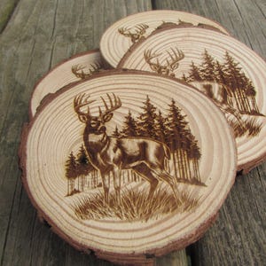 May include: A set of four wooden coasters with a deer and forest scene etched into the surface. The coasters are made from natural wood slices, featuring a deer standing in a grassy area with a forest in the background. The coasters are on a wooden surface.