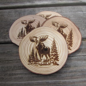 May include: Set of four wooden coasters featuring a moose design. Each coaster is made of natural wood and has a unique grain pattern. The moose is burned into the wood and is surrounded by trees.
