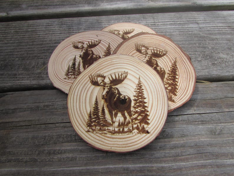 May include: Set of four wooden coasters featuring a moose design. Each coaster is made of natural wood and has a unique grain pattern. The moose is burned into the wood and is surrounded by trees.
