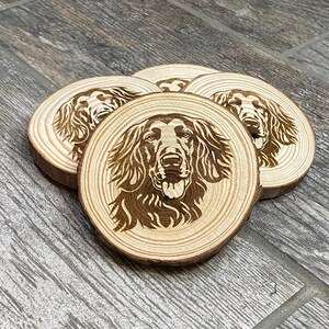 May include: Set of four wooden coasters with a laser-etched design of a dog's face. The coasters are round, made from natural wood, and feature a detailed portrait of a dog breed. The wood has a light brown color, and the dog's image is a darker brown.
