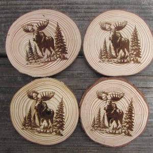 May include: Set of four wooden coasters featuring a moose silhouette design with a forest background. The coasters are circular and have a natural wood finish.