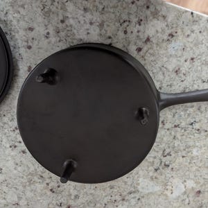 May include: A black cast iron skillet with a long handle and three small feet. The skillet is round and has a smooth surface. The handle has a loop at the end for hanging.