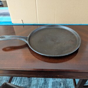 May include: A round, flat cast iron griddle with a short, integrated handle. The surface is dark, textured, and matte, featuring a low-profile rim around the edge. The handle has a small hole at the end for hanging, and the overall construction is solid.