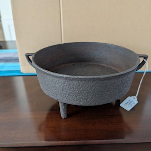 May include: A round, dark gray cast iron pot with a textured exterior, three short legs, and two small side handles. A white string with a small rectangular tag is attached to one handle, featuring handwritten text that is partially visible.