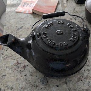 May include: A black cast iron kettle with a handle and spout. The lid is embossed with the words "VO CO-OP BONE OS". The kettle is sitting on a speckled countertop.