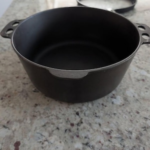 May include: A black cast iron Dutch oven with two handles, each with two holes. The interior is dark, and the rim has a silver-colored edge. The pot sits on a speckled countertop.