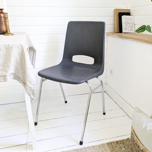 May include: A dark grey plastic chair with a curved back and a rectangular cut-out. The chair has four chrome legs with black rubber feet. The chair is in a room with white walls and a white floor.
