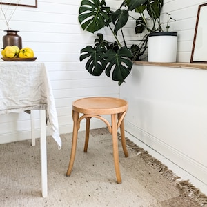 Op de afbeelding: Een lichte houten kruk met een ronde zitting en vier poten. De kruk staat in een kamer met witte muren en een lichtbruin tapijt.
