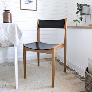 Könnte beinhalten: Ein Holzstuhl mit schwarzem Sitz und Rückenlehne. Der Stuhl hat ein hellbraunes Gestell und ein geschwungenes Sitzdesign. Der Stuhl steht auf einem beigen Teppich in einem Raum mit weißen Wänden.