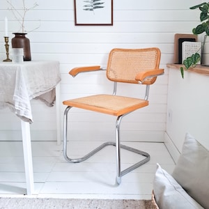 Op de afbeelding: Een mid-century modern stoel met een geweven rieten zitting en rugleuning. De stoel heeft een licht houten frame met armleuningen en een verchroomd stalen onderstel. De stoel staat tegen een witte muur en vloer.