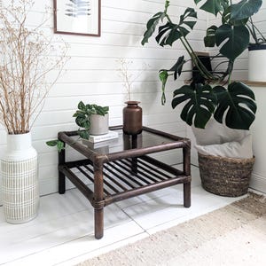 Könnte beinhalten: Ein dunkelbrauner, quadratischer Couchtisch mit Glasplatte und Lattenrost. Der Tisch ist von dekorativen Gegenständen umgeben, darunter eine große weiße Vase, Topfpflanzen und ein Weidenkorb. Der Raum hat weiße Wände und einen neutralen Teppich.