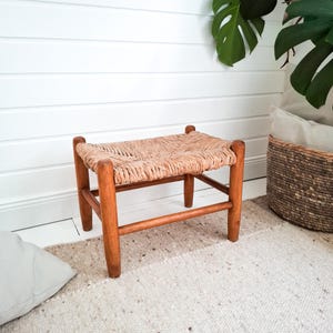 Puede incluir: Un pequeño taburete con asiento tejido y patas de madera. El asiento es de fibras naturales y tiene forma rectangular. Las patas son de madera de color marrón claro. El taburete está colocado sobre una alfombra texturizada, mide unos 40 cm de largo.