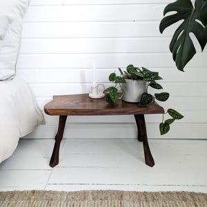 Peut inclure: Un tabouret en bois avec une finition en bois naturel et un plateau à bord vif. Le tabouret a trois pieds et est posé sur un sol blanc. Une plante en pot avec des feuilles vertes est posée sur le tabouret.