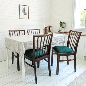 Könnte beinhalten: Ein Esszimmer-Set mit einem weißen Tisch mit einer Leinentischdecke und vier Holzstühlen. Die Stühle haben dunkle Holzrahmen und smaragdgrüne Samtsitzkissen. Ein gerahmter Farn-Druck hängt an der weißen Wand.