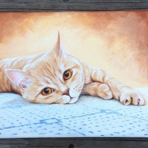 Op de afbeelding: Een schilderij van een gemberkat die op een blauw-witte deken ligt. De kat heeft heldere gele ogen en kijkt recht naar de kijker.