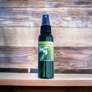 May include: A green glass bottle with a black spray top and a white label that says "Sandy Land Studio Moisturizing Mist Lily of the Valley". The label features a watercolour illustration of lily of the valley flowers.