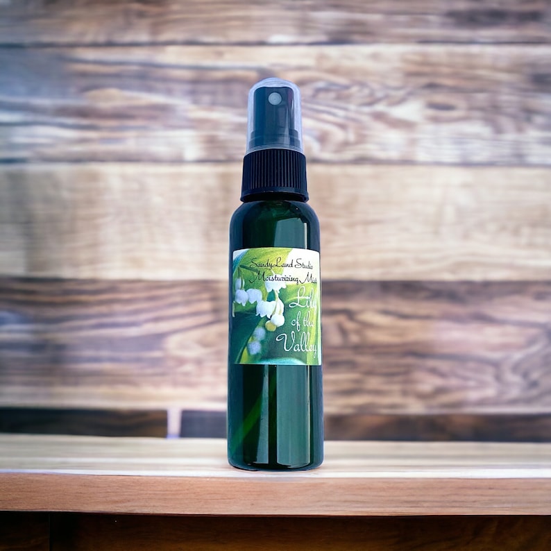 May include: A green glass bottle with a black spray top and a white label that says "Sandy Land Studio Moisturizing Mist Lily of the Valley". The label features a watercolour illustration of lily of the valley flowers.