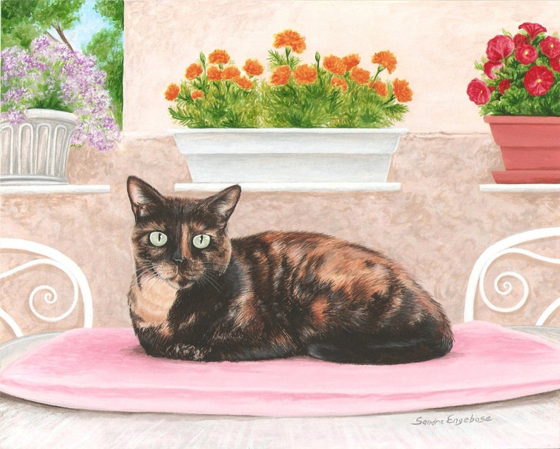 Op de afbeelding: Een schildpadkat met groene ogen ligt op een roze kussen op een witte patiotafel. De kat kijkt recht naar de kijker. Er zijn potplanten met oranje bloemen op de achtergrond.