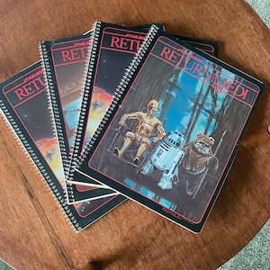Può includere: Quattro quaderni a spirale con copertine nere e testo bianco. Le copertine presentano immagini del film di Star Wars Il Ritorno dello Jedi. Il testo sulle copertine recita "Star Wars Il Ritorno dello Jedi".