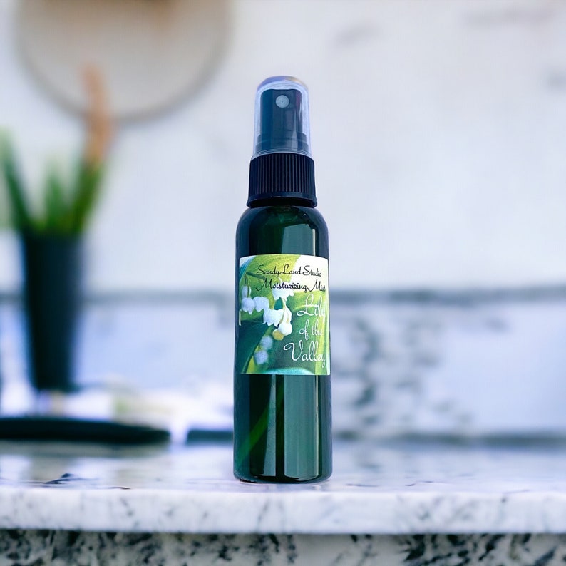 May include: A green glass bottle with a black spray top and a white label that says "Sandy Land Studio Moisturizing Mist Lily of the Valley". The label features a watercolour illustration of lily of the valley flowers.