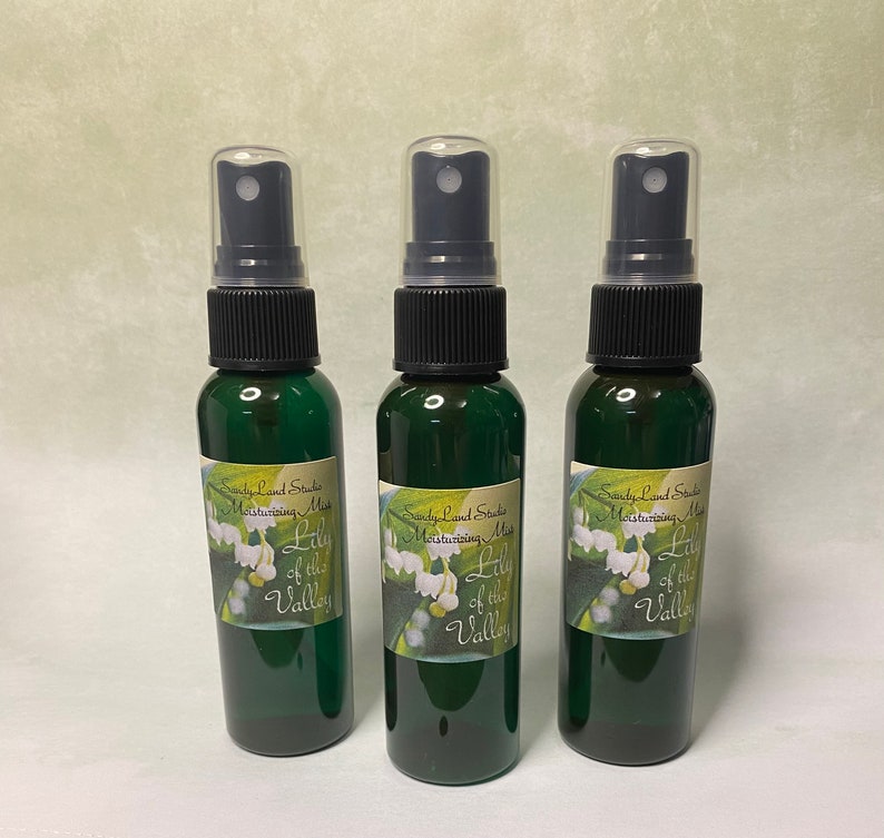 May include: Three green glass bottles with black spray tops and white labels. The labels say "Sandy Land Studio Moisturizing Mist Lily of the Valley".