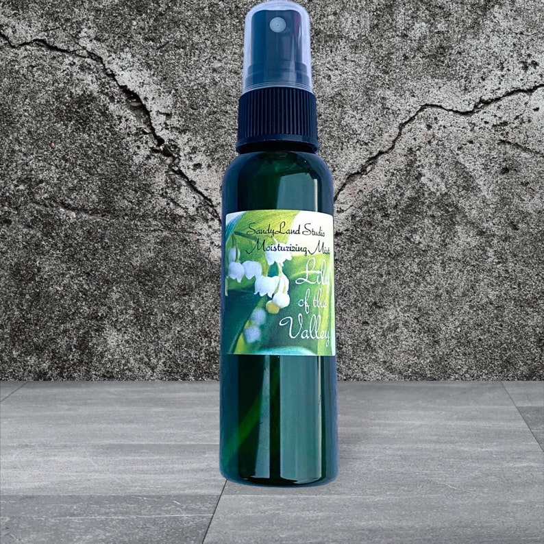 May include: A green glass bottle with a black spray top and a white label that says "SandyLand Studio Moisturizing Mist Lily of the Valley". The label has a picture of lily of the valley flowers.
