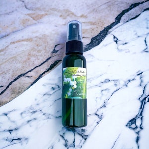 May include: A green glass bottle with a black spray top and a white label that says "Sandy Land Studio Moisturizing Mist Lily of the Valley". The bottle is sitting on a white and grey marble surface.