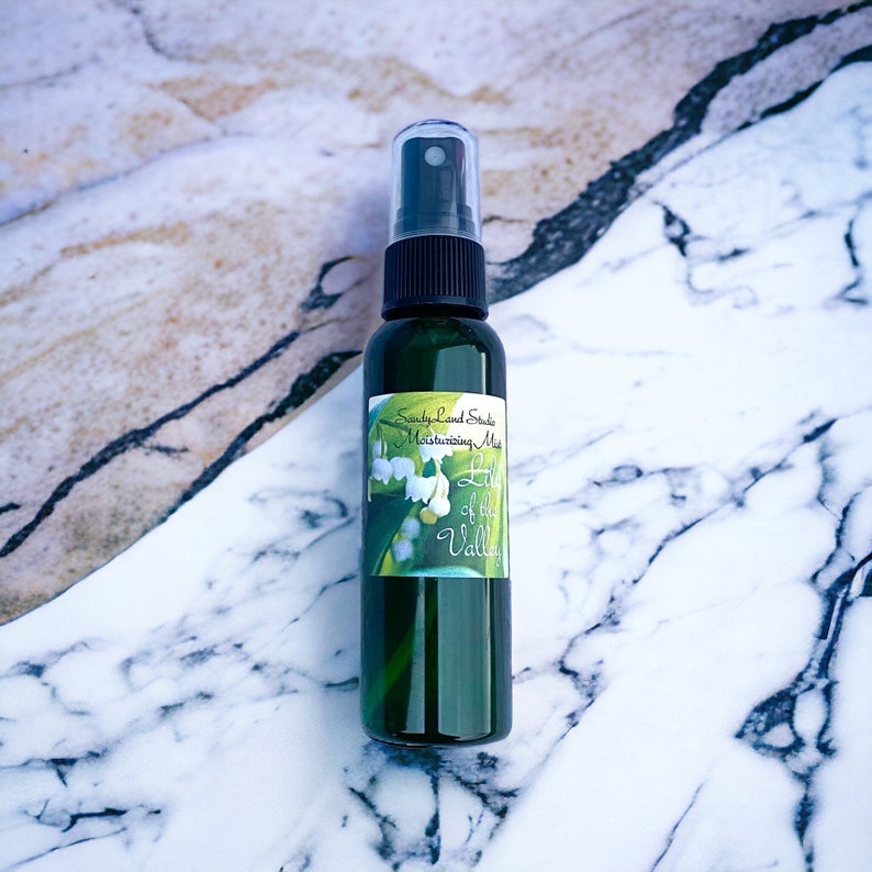 May include: A green glass bottle with a black spray top and a white label that says "Sandy Land Studio Moisturizing Mist Lily of the Valley". The bottle is sitting on a white and grey marble surface.