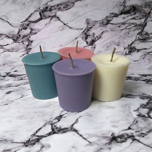 Op de afbeelding: Vier theelichtjes in verschillende kleuren: teal, lavendel, crème en roze. De kaarsen zijn in een halve cirkel op een wit marmeren oppervlak gerangschikt.