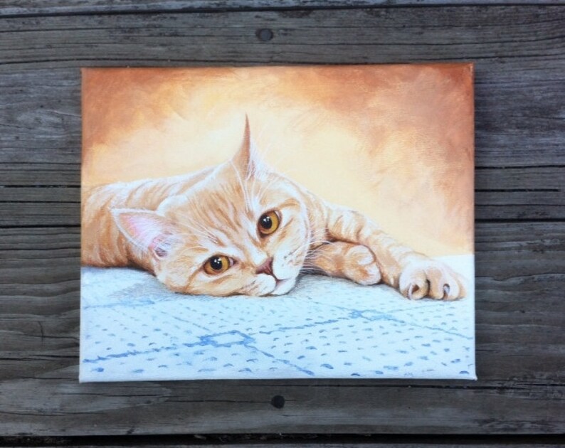 Op de afbeelding: Een schilderij van een gemberkat die op een blauw-witte deken ligt. De kat heeft heldere gele ogen en kijkt recht naar de kijker.