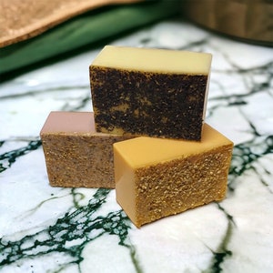 May include: Four handmade soap bars in various colors are stacked. The top bar is light yellow, the second is dark brown with visible seeds, the third is light pink, and the bottom bar is tan with a textured surface. The soaps are on a marble surface.