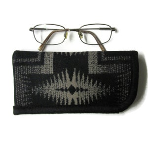 May include: Eyeglasses with a gold-colored frame and brown temples rest on top of a black and gray patterned glasses case. The case has a geometric design and a black border, providing a stylish storage solution for eyewear.