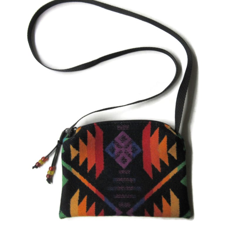 Puede incluir: Un peque&ntilde;o bolso bandolera negro con un colorido estampado geom&eacute;trico en tonos rojos, naranjas, amarillos, verdes y morados. El bolso tiene una correa negra y un cierre de cremallera con detalles de cuentas. El bolso mide aproximadamente 18 cm de ancho.
