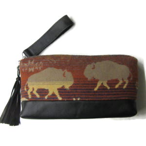 May include: A rectangular clutch with a black leather base and wrist strap. The upper portion features a patterned fabric in shades of orange, brown, and blue, with two tan bison silhouettes. A black tassel hangs from the zipper.