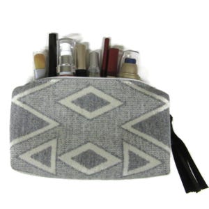 May include: A gray cosmetic bag with a geometric diamond pattern in white. The bag is filled with various makeup items, including brushes, lipsticks, and bottles. A black tassel hangs from the zipper. The bag is designed to hold beauty products.