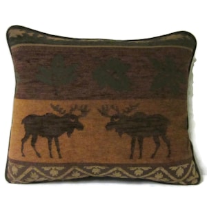 May include: Rectangular decorative pillow with a brown and tan color scheme. The pillow features a repeating pattern of moose silhouettes and leaf designs. The top and bottom borders have a leaf pattern, and the center has two moose.