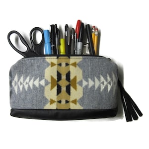 May include: A gray pencil case with a geometric pattern in white, tan, and black, filled with pens, pencils, and scissors. The bottom of the case is black, and it has a black tassel zipper pull.