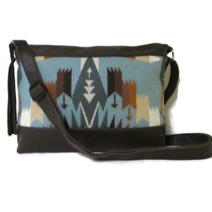 May include: A rectangular crossbody bag with a geometric pattern in shades of blue, brown, and cream. The bag has a dark brown leather base, trim, and strap. The strap is adjustable and the bag has a tassel.