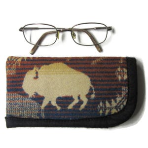 May include: A pair of silver-toned metal eyeglasses with thin frames positioned above a rectangular fabric eyeglass case. The case features a woven pattern of horizontal blue and rust-colored stripes with a large cream-colored buffalo silhouette.