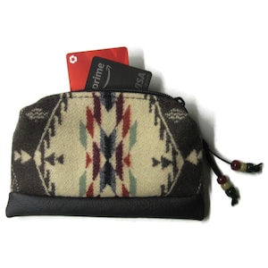 May include: A small, zippered wallet with a Southwestern pattern in cream, brown, red, and blue. The bottom is black leather. Two cards are visible inside. A leather pull with decorative beads hangs from the zipper.