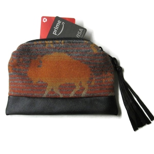 May include: A small, zippered wallet with a bison design. The wallet features a black leather base and a patterned fabric top in orange, gray, and red. A black tassel hangs from the zipper pull. A red Prime card and a black Visa card are visible.