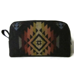 May include: A rectangular zippered pouch featuring a black base with a central geometric pattern in shades of tan, rust, and muted blue. The design consists of stacked diamonds and triangular shapes, and the pouch includes a small side carry loop.