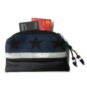 May include: A small, zippered wallet with a navy blue top section featuring three black stars. A white stripe separates the navy blue section from the black leather bottom. Two credit cards are visible inside. The wallet has a black zipper and a beaded pull.