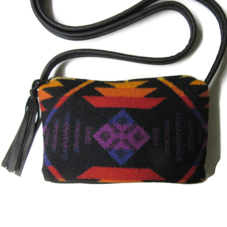 May include: A rectangular crossbody bag with a black background and a colourful geometric pattern in orange, red, purple, and blue. The bag has a black strap and a black tassel. The bag is made of a soft, woven material.