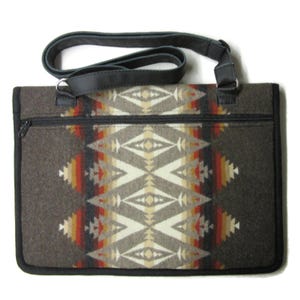 May include: A rectangular tote bag with a Southwestern geometric pattern in shades of brown, beige, red, and orange. The bag has a black zipper, black leather trim, and a black leather shoulder strap. The bag is made of a thick, woven material.