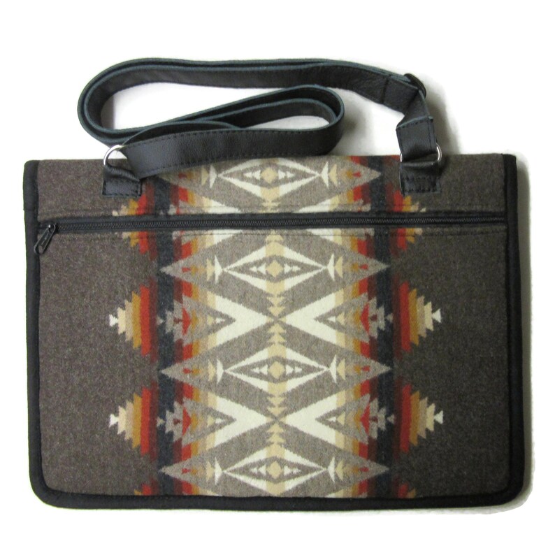 May include: A rectangular tote bag with a Southwestern geometric pattern in shades of brown, beige, red, and orange. The bag has a black zipper, black leather trim, and a black leather shoulder strap. The bag is made of a thick, woven material.