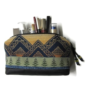 May include: A zippered pouch featuring a woven pattern of dark blue mountains over a light green band with small pine tree motifs. The base is solid black material. The pouch is filled with various cosmetic items including lipsticks and bottles.