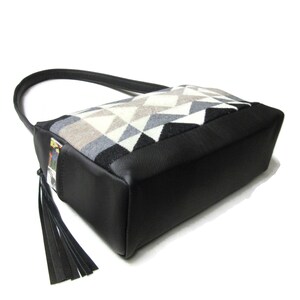 May include: A black and geometric patterned handbag with a black leather base and handle. The bag features a geometric pattern in shades of gray, white, and black. A black tassel hangs from the side.