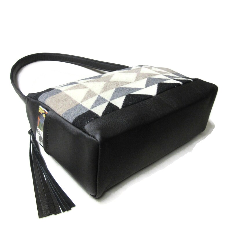 May include: A black and geometric patterned handbag with a black leather base and handle. The bag features a geometric pattern in shades of gray, white, and black. A black tassel hangs from the side.